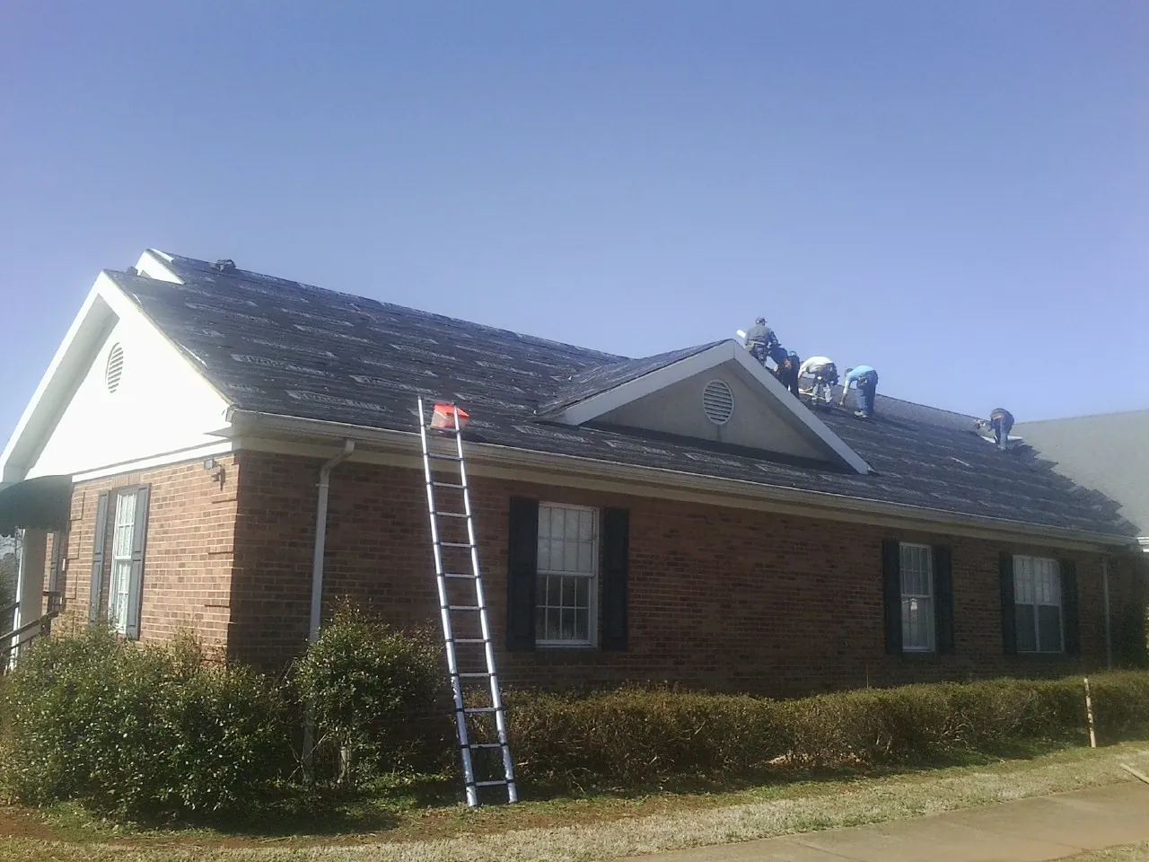 Crew tearing shingles off a brick ranch roof with a ladder set — Metal Roof Repair work by our crew in Geneva